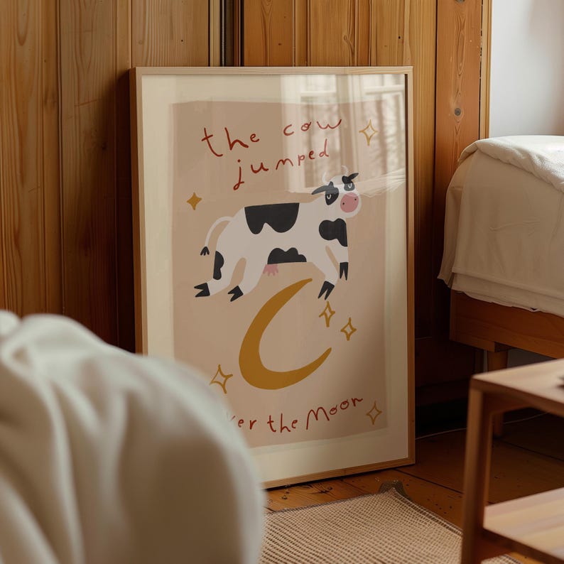 May include: Framed art print featuring a whimsical illustration of a cow jumping over a yellow crescent moon. The text "the cow jumped over the moon" is written in red and black. The artwork has a light beige background and is surrounded by a wooden frame.