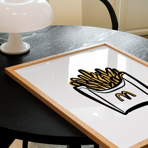 K&ouml;nnte beinhalten: Ein gerahmter Druck einer gelben und schwarzen Illustration einer McDonald's-Pommes-Frites-Schachtel mit dem McDonald's-Logo an der Seite. Der Druck ist auf wei&szlig;em Hintergrund und in einem hellbraunen Holzrahmen gerahmt.
