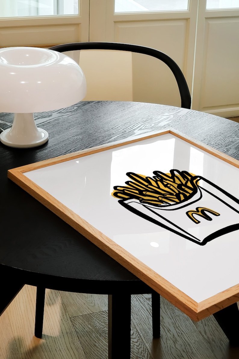 K&ouml;nnte beinhalten: Ein gerahmter Druck einer gelben und schwarzen Illustration einer McDonald's-Pommes-Frites-Schachtel mit dem McDonald's-Logo an der Seite. Der Druck ist auf wei&szlig;em Hintergrund und in einem hellbraunen Holzrahmen gerahmt.