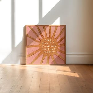 May include: A framed square artwork with a pink sunburst design and gold rays. The white text reads "AND ALL IT TOOK WAS ANOTHER SUNRISE". The artwork is displayed on a light wooden floor, with a shadow cast across it.