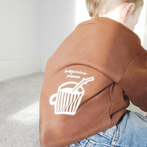 May include: A brown sweatshirt with the text "babyccino please" and a graphic of a coffee cup with a straw. The sweatshirt is paired with blue denim jeans. The image is taken from behind.