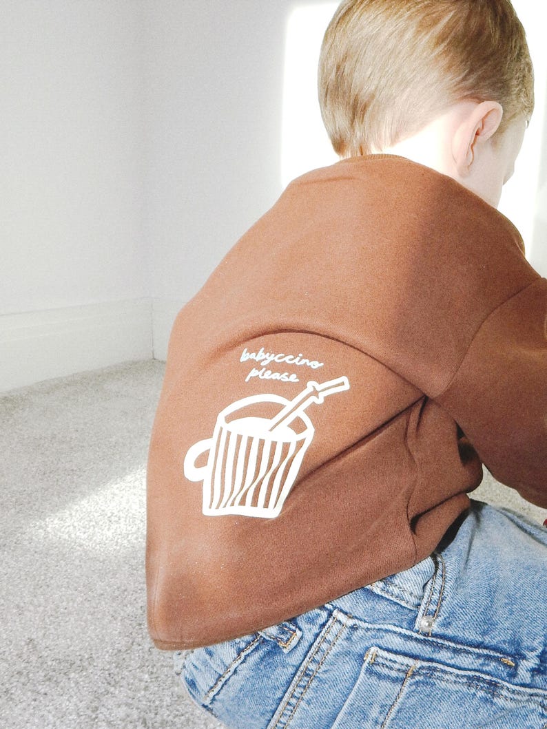 May include: A brown sweatshirt with the text "babyccino please" and a graphic of a coffee cup with a straw. The sweatshirt is paired with blue denim jeans. The image is taken from behind.