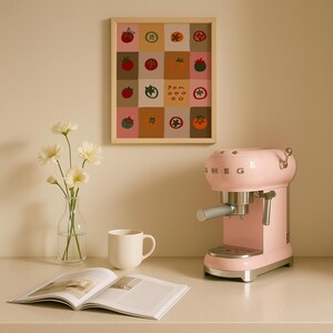 May include: A light pink SMEG espresso machine sits on a white countertop next to a white mug and an open magazine. A framed print with tomato illustrations hangs on the wall. A vase of white flowers is also on the counter.
