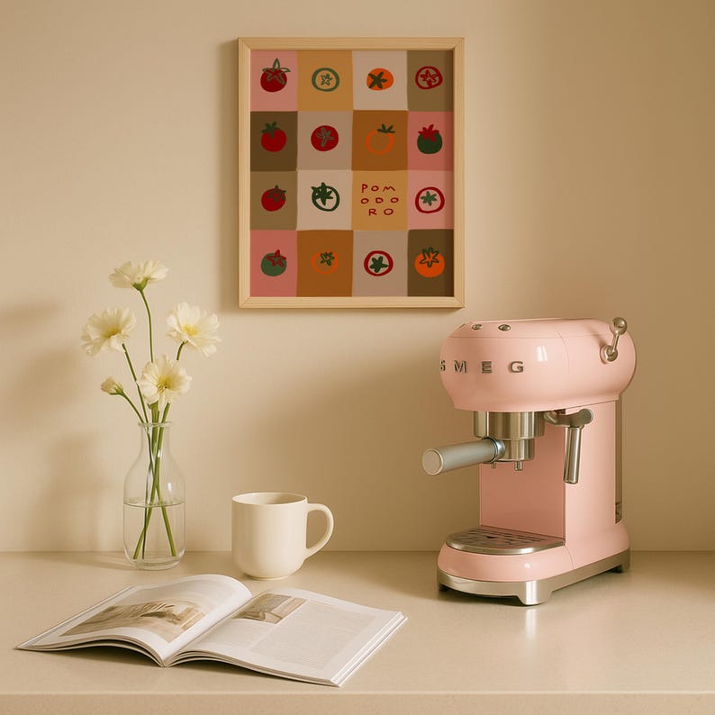 May include: A light pink SMEG espresso machine sits on a white countertop next to a white mug and an open magazine. A framed print with tomato illustrations hangs on the wall. A vase of white flowers is also on the counter.