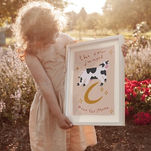 May include: A framed art print featuring a whimsical illustration of a cow jumping over a crescent moon. The text reads "the cow jumped over the moon." The artwork has a light beige background with a white frame. The cow is black and white.