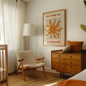 May include: A framed poster with the text "YOU ARE MY SUNSHINE" and a smiling sun illustration hangs on a white wall. A mid-century modern style wooden dresser and chair are in the room. A floor lamp and a crib are also visible.