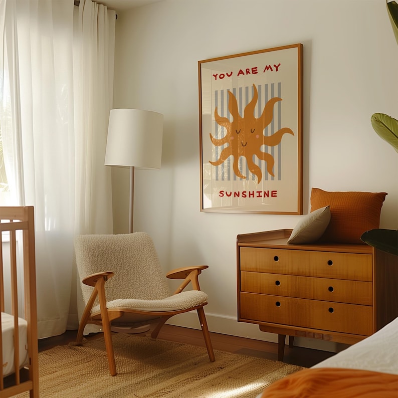 May include: A framed poster with the text "YOU ARE MY SUNSHINE" and a smiling sun illustration hangs on a white wall. A mid-century modern style wooden dresser and chair are in the room. A floor lamp and a crib are also visible.