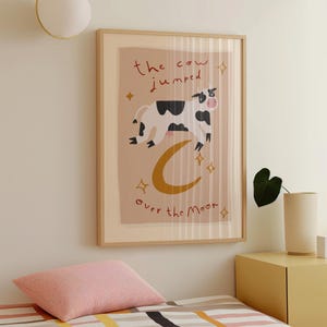 May include: A framed art print featuring a whimsical illustration of a cow jumping over a crescent moon. The cow is black and white, and the text reads "the cow jumped over the moon". The artwork has a light brown background with gold stars.