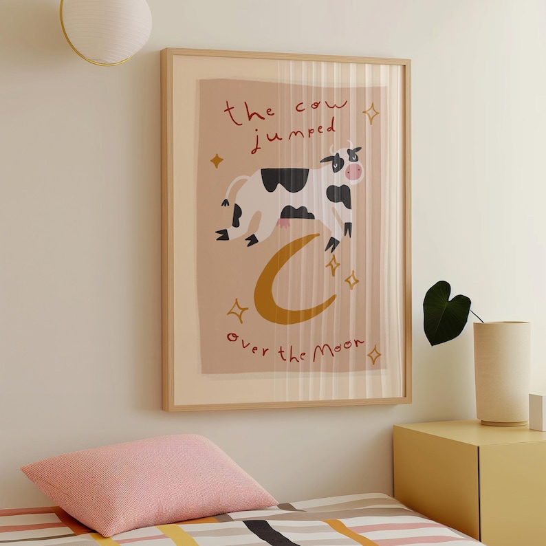 May include: A framed art print featuring a whimsical illustration of a cow jumping over a crescent moon. The cow is black and white, and the text reads "the cow jumped over the moon". The artwork has a light brown background with gold stars.