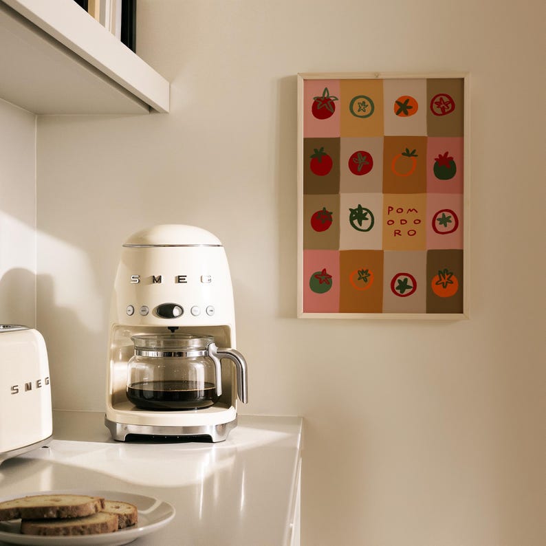 May include: Cream-colored SMEG coffee maker with a glass carafe filled with coffee. A framed artwork with tomato illustrations hangs on the wall. The artwork has the word "Pomodoro" in the center. A SMEG toaster and a plate of toast are also visible.