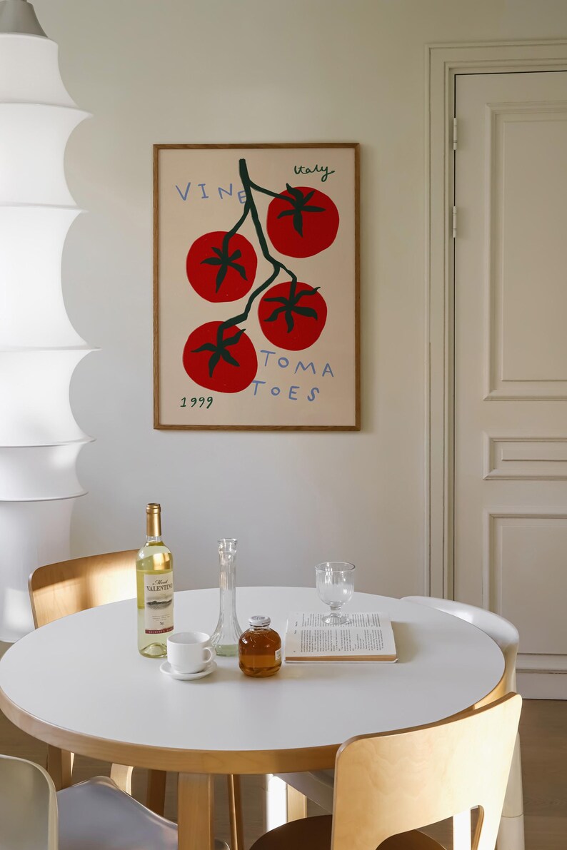May include: A framed illustration of a tomato vine with four red tomatoes and the text "VINE ITALY TOMATOES 1999". The illustration is on a white background and is hanging on a white wall. A white table with a bottle of wine, a glass, a vase, and a book is in the foreground.