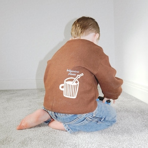 May include: A brown sweatshirt with the text "babyccino please" and a white graphic of a coffee cup with a straw. The sweatshirt is worn with blue denim jeans. The child is kneeling on a gray carpet.