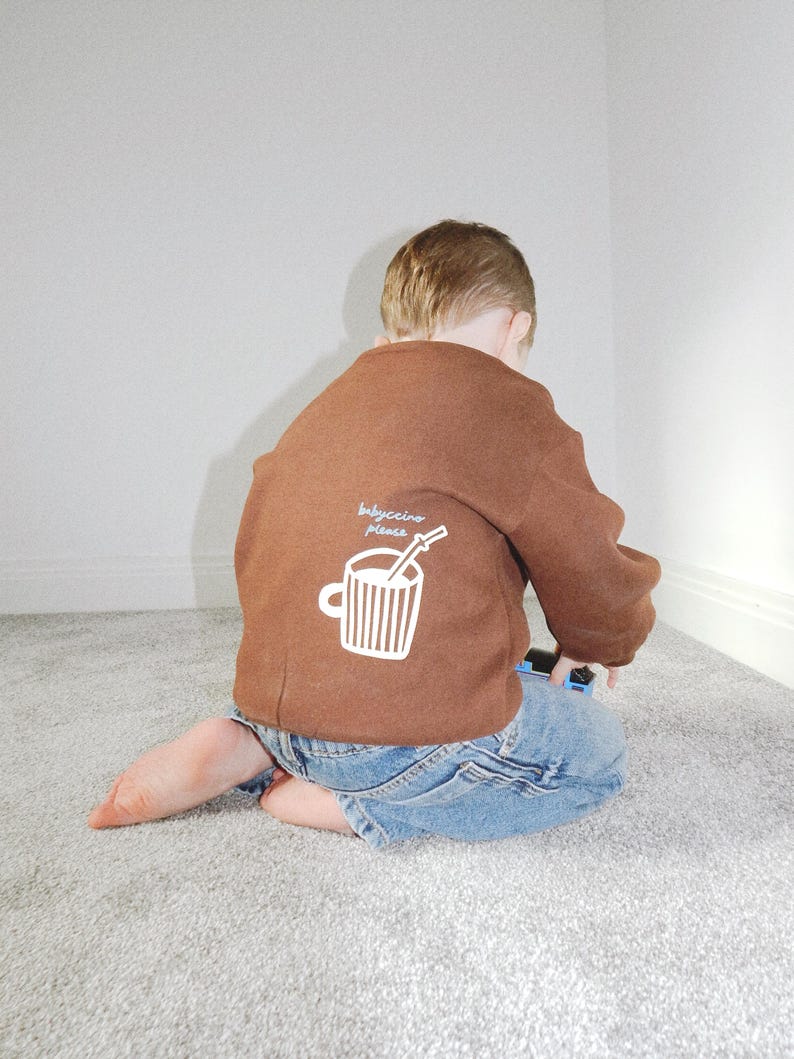 May include: A brown sweatshirt with the text "babyccino please" and a white graphic of a coffee cup with a straw. The sweatshirt is worn with blue denim jeans. The child is kneeling on a gray carpet.