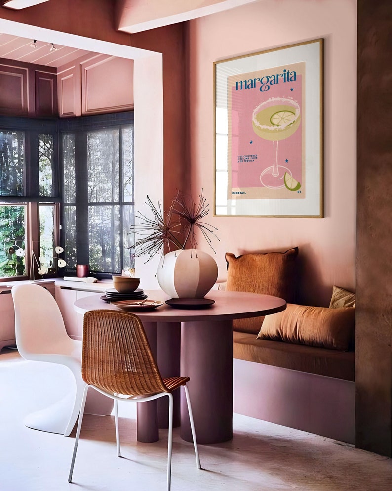 Peut inclure: Une affiche encadr&eacute;e rose et or avec le mot "Margarita" en bleu. L'affiche repr&eacute;sente un cocktail margarita rose avec un quartier de citron vert et un motif d'&eacute;toiles. L'affiche est accroch&eacute;e &agrave; un mur rose dans une salle &agrave; manger avec une table ronde et des chaises.