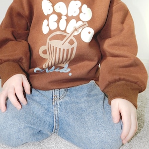 May include: A brown sweatshirt with the words "Baby Cino Club" in white bubble letters and a graphic of a coffee cup with a straw. The sweatshirt has brown ribbed cuffs and a relaxed fit. The child is wearing blue jeans.