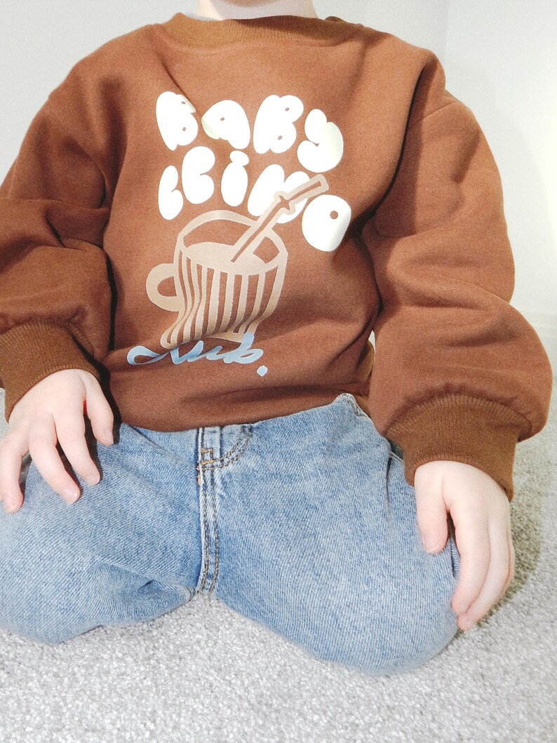 May include: A brown sweatshirt with the words "Baby Cino Club" in white bubble letters and a graphic of a coffee cup with a straw. The sweatshirt has brown ribbed cuffs and a relaxed fit. The child is wearing blue jeans.