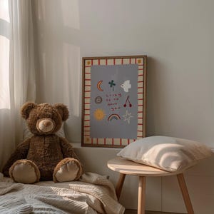 May include: A brown teddy bear sits on a beige blanket in front of a framed print with a light blue background and red and white stripes. The print features a variety of colorful illustrations, including a sun, a rainbow, a star, a cherry, a bird, a clover, and a crescent moon. The text on the print reads "Lucky is to have you".