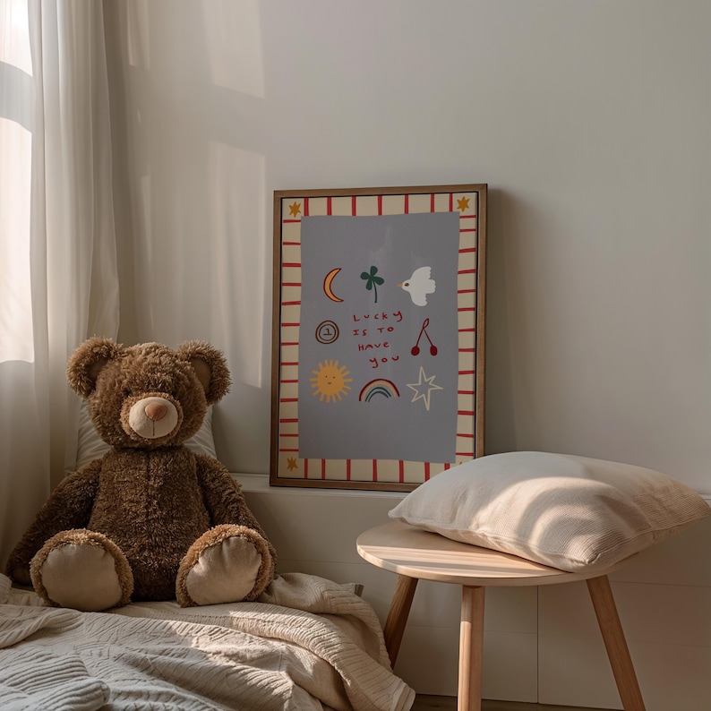 May include: A brown teddy bear sits on a beige blanket in front of a framed print with a light blue background and red and white stripes. The print features a variety of colorful illustrations, including a sun, a rainbow, a star, a cherry, a bird, a clover, and a crescent moon. The text on the print reads "Lucky is to have you".