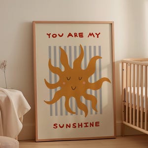 May include: A framed art print featuring a smiling sun illustration in shades of orange and yellow. The print has the words "YOU ARE MY SUNSHINE" in red. The background has vertical gray and white stripes.