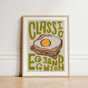 May include: A framed print with a light brown background. The print features a cartoon illustration of a sandwich with an egg inside. The text on the print reads 