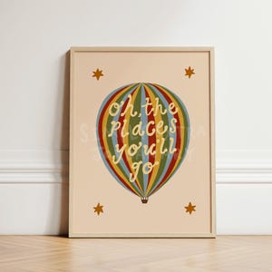 May include: A framed wall art print on a light beige background featuring a multi-coloured striped hot air balloon with the text Oh, the places you'll go written in white script across the centre, surrounded by four small orange stars.
