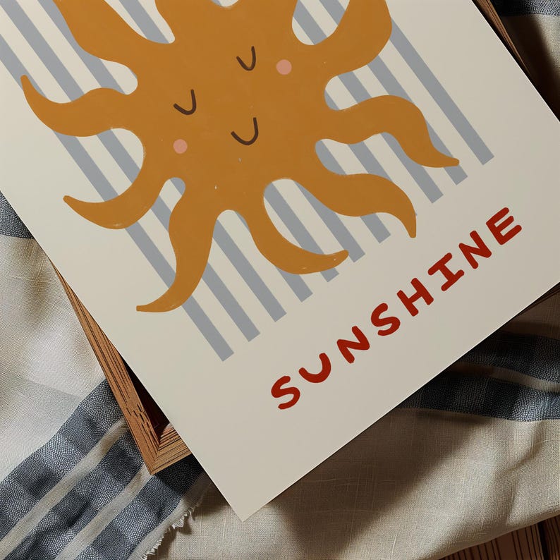 May include: A framed print featuring a cartoon sun in shades of orange and peach, with a smiling face. The background has light blue and gray stripes. The word "SUNSHINE" is printed in red at the bottom.