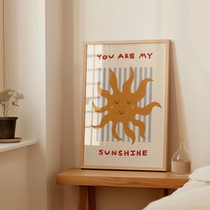 May include: A framed art print featuring a smiling sun illustration in shades of orange and yellow. The sun is set against a backdrop of vertical blue and white stripes. The words "YOU ARE MY SUNSHINE" are printed in red.