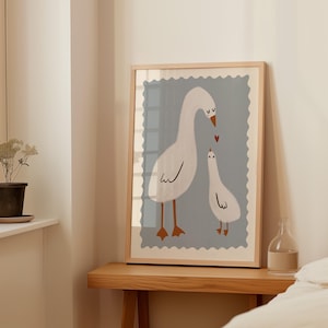K&ouml;nnte beinhalten: Gerahmter Kunstdruck mit einer Illustration von zwei wei&szlig;en G&auml;nsen mit braunen Beinen und Schn&auml;beln. Die G&auml;nse sind vor einem hellblauen Hintergrund mit dekorativem Rand platziert. Ein kleines rotes Herz befindet sich zwischen den G&auml;nsen.
