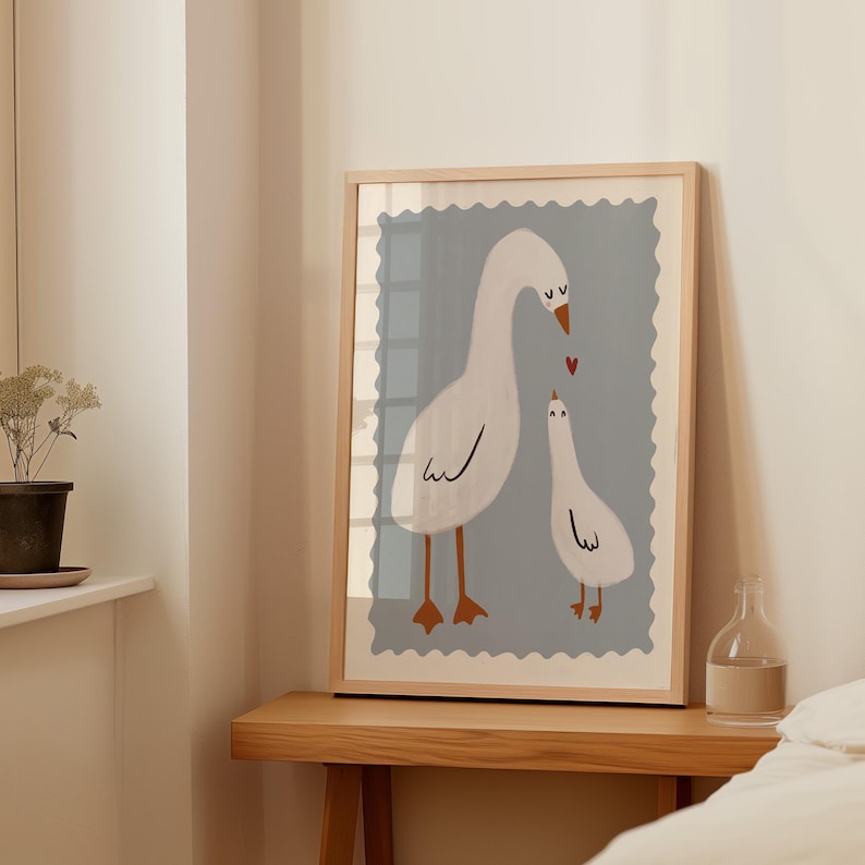 K&ouml;nnte beinhalten: Gerahmter Kunstdruck mit einer Illustration von zwei wei&szlig;en G&auml;nsen mit braunen Beinen und Schn&auml;beln. Die G&auml;nse sind vor einem hellblauen Hintergrund mit dekorativem Rand platziert. Ein kleines rotes Herz befindet sich zwischen den G&auml;nsen.