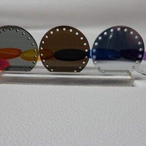 May include: Three round, semi-transparent acrylic pieces in shades of gray, brown, and blue, each with evenly spaced holes around the edge. They are displayed on a clear acrylic stand. Other colorful acrylic shapes are visible in the background.