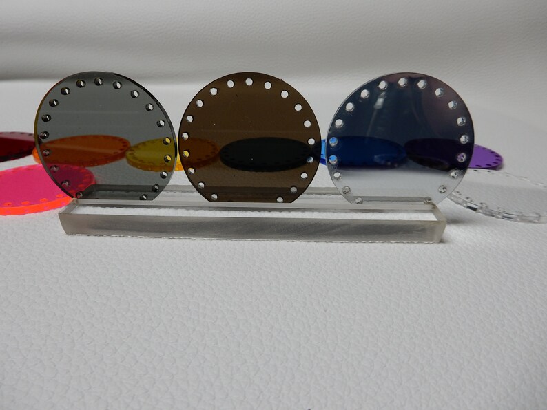 May include: Three round, semi-transparent acrylic pieces in shades of gray, brown, and blue, each with evenly spaced holes around the edge. They are displayed on a clear acrylic stand. Other colorful acrylic shapes are visible in the background.