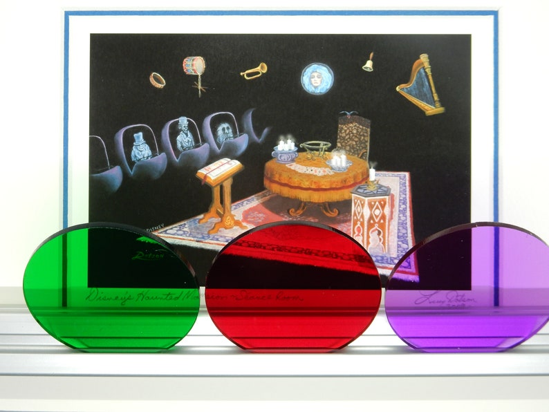 May include: Three colored, oval-shaped glass pieces in green, red, and purple, with a framed illustration of a haunted mansion scene. The green glass has the text "Disney's Haunted Mansion". The red glass has the text "Leota's Room".