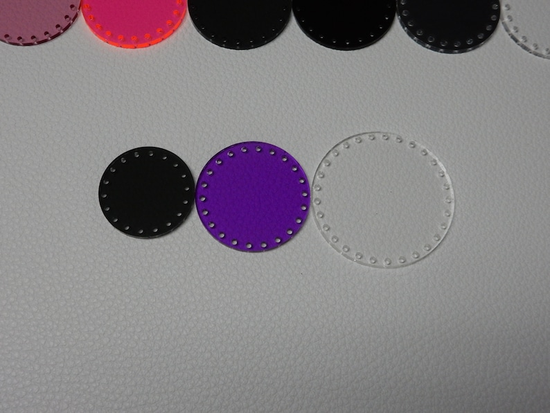 May include: A collection of circular craft pieces in various colors. The pieces include black, purple, and clear acrylic circles, each with evenly spaced holes around the perimeter. Other colors include pink and orange.