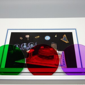 May include: An artwork featuring a framed print with a surrealist scene, partially obscured by three colored, circular filters: green, red, and purple. The print depicts floating objects and figures against a dark background.