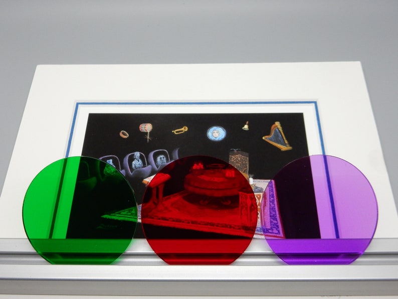 May include: An artwork featuring a framed print with a surrealist scene, partially obscured by three colored, circular filters: green, red, and purple. The print depicts floating objects and figures against a dark background.