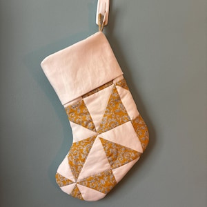 Puede incluir: Un calcetín navideño acolchado con un patrón geométrico de triángulos blancos sólidos alternados con triángulos de color amarillo mostaza con un estampado floral blanco. El calcetín tiene un puño blanco liso en la parte superior y una pequeña lazada.