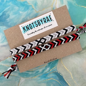 May include: Two friendship bracelets on a brown card with the text "KnotsByRae Friendship Bracelets for all occasions" and "KnotsByRae" at the bottom. One bracelet is black, white, and red with a chevron pattern. The other bracelet is grey, white, and black with a chevron pattern.