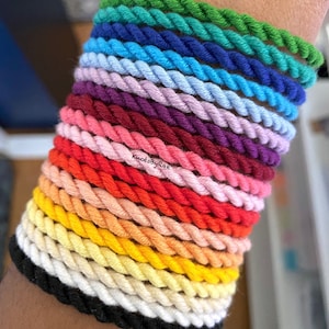 May include: A stack of 18 colourful braided rope bracelets in various shades of green, blue, purple, pink, red, orange, yellow, and white. The bracelets are stacked on a person's wrist. The text "KnotsByRae" is visible on the bracelets.