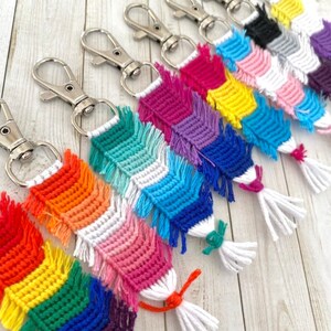 May include: A set of eight colourful crocheted keychains with fringe tassels. Each keychain features a different colour pattern, including rainbow, pink and white, blue and white, yellow and blue, purple and blue, red and white, black and white, and grey and white.
