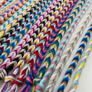 May include: A collection of colourful friendship bracelets with various patterns and colours, including red, blue, green, yellow, white, and pink. The bracelets are made with a braided technique and have a knotted finish.