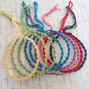 May include: A collection of colourful friendship bracelets made with twisted cord. The bracelets are in various colours, including yellow, green, blue, purple, red, and black. The bracelets are arranged in a circle, with the ends of the cords tied together.