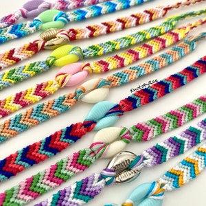 May include: Close-up of several colourful friendship bracelets with chevron patterns. Each bracelet features a seashell accent in various colours, including yellow, pink, and blue. The bracelets are handmade and have a bohemian style.