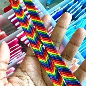 Peut inclure: Deux bracelets d'amitié en chevron arc-en-ciel faits avec du fil à broder coloré. Les bracelets sont tenus dans une main.