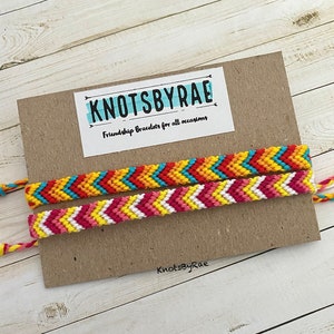 May include: Two chevron-patterned friendship bracelets in vibrant colours are shown on a cardboard backing. One bracelet has red, yellow, and blue, the other pink, yellow, and white. The card reads "KNOTSBYRAE" and "Friendship Bracelets for all occasions."