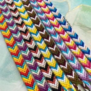 May include: A set of eight colourful friendship bracelets with chevron patterns. The bracelets are made with different coloured embroidery floss and feature a variety of colour combinations, including red, orange, yellow, green, blue, purple, and white.