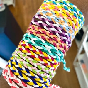 May include: A stack of colourful friendship bracelets made with braided string. The bracelets are in various colours, including pink, blue, green, yellow, and white.