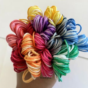 May include: A collection of embroidery floss spools in a rainbow of colours, including red, orange, yellow, green, blue, purple, and white. The spools are arranged in a circular pattern and are tied together with a white string. The text "KnotsByRae" is visible at the bottom of the image.