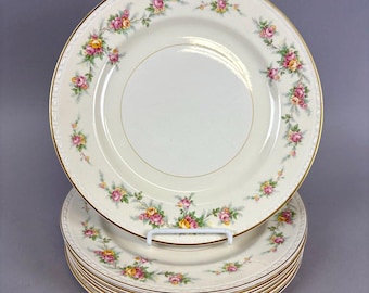 May include: A stack of cream-colored plates with a floral rose pattern in pink, yellow, and green. Each plate has a gold rim and a gold inner ring. The plates are stacked on a white plate stand.