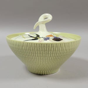 May include: A pale green ceramic bowl with a lid. The bowl has a textured, vertical striped pattern. The lid features a decorative design with black, blue, orange, and brown elements. A white, looped handle sits atop the lid.
