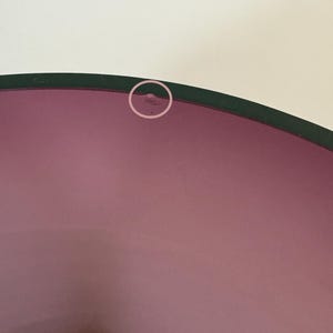 May include: A close-up of a purple glass bowl with a dark green rim. The bowl has a smooth, glossy surface and a slight imperfection on the rim. The background is a plain white.